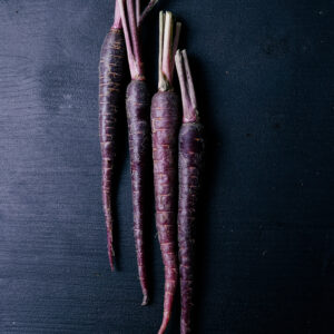 four long, deep purple carrots laid vertically on a dark wooden table