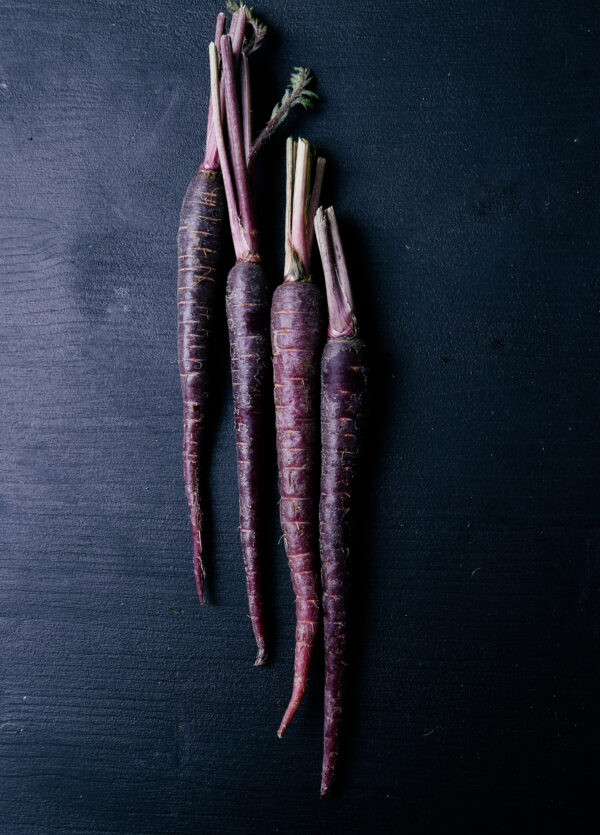 four long, deep purple carrots laid vertically on a dark wooden table
